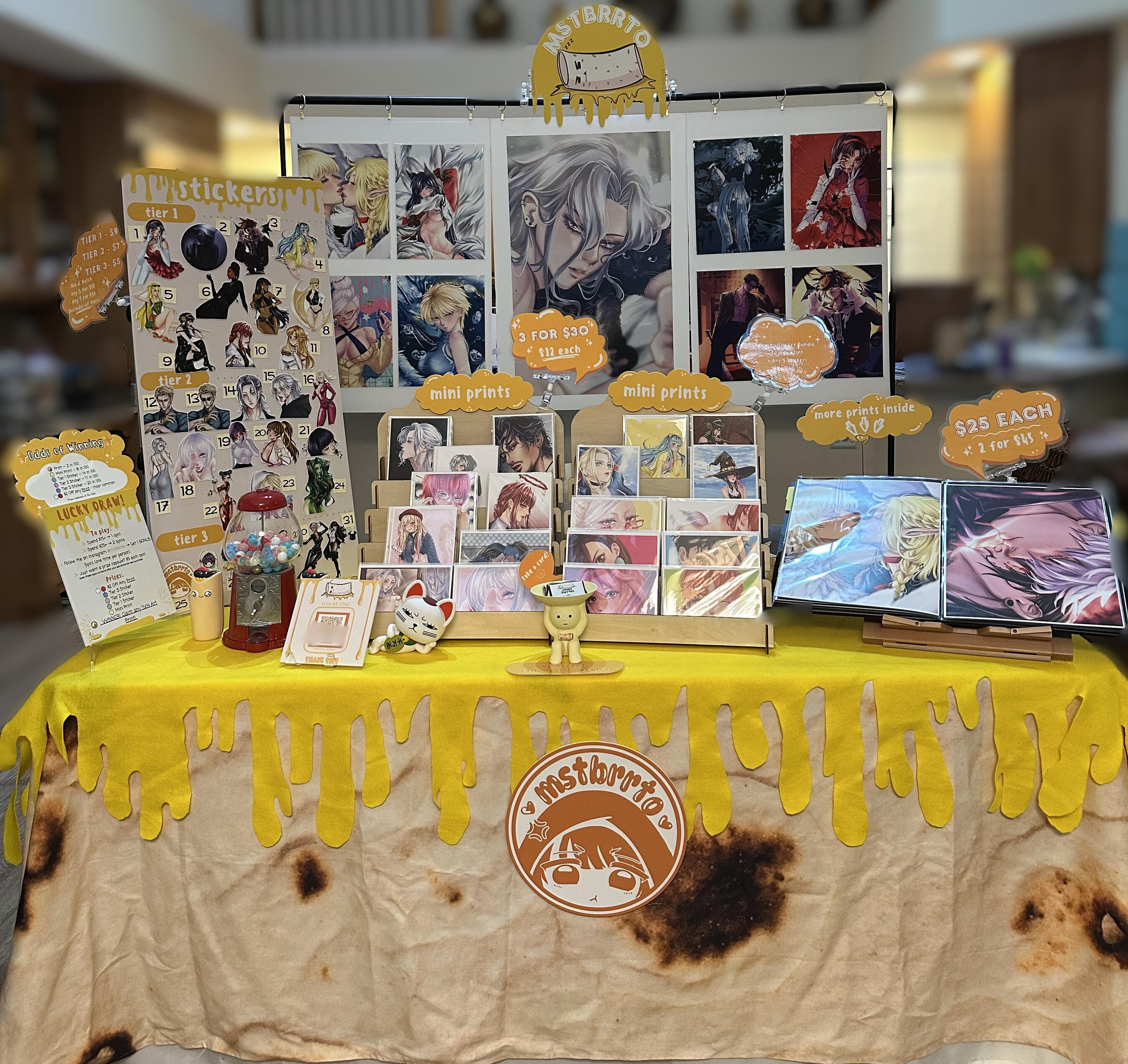 mstbrrto Artist Alley Setup mstbrrto convention booth featuring prints, stickers, and yellow honey‑drip tablecloth.